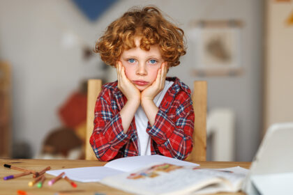 Kind met rode krullen van ongeveer 6 jaar oud zit met handen onder de kin, staart voor zich uit met helder blauwe ogen en huiswerk ligt op tafel.
